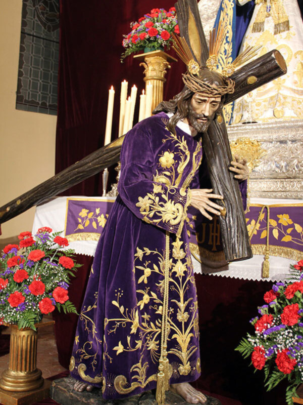 Estatua de Cristo en la Sagrada Familia, vestida con una túnica púrpura con bordados dorados, sosteniendo un cetro y rodeada por flores y velas en una iglesia.