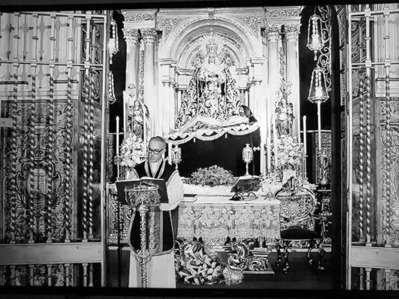 En una iglesia, un sacerdote con un hábito negro y gafas se encuentra frente a una araña de altar adornada con flores. La imagen captura la solemnidad y el respeto en un entorno de culto religioso.