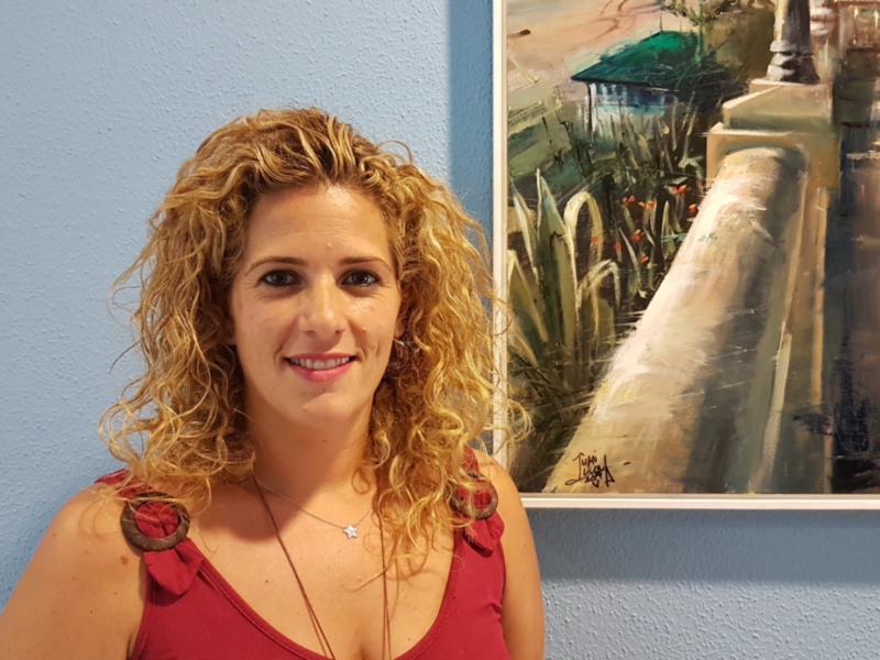 Una mujer con cabello rizado se encuentra frente a una pared azul, sonriendo. A su lado derecha, hay un cuadro de paisaje pintado en tonos de verde y beige.