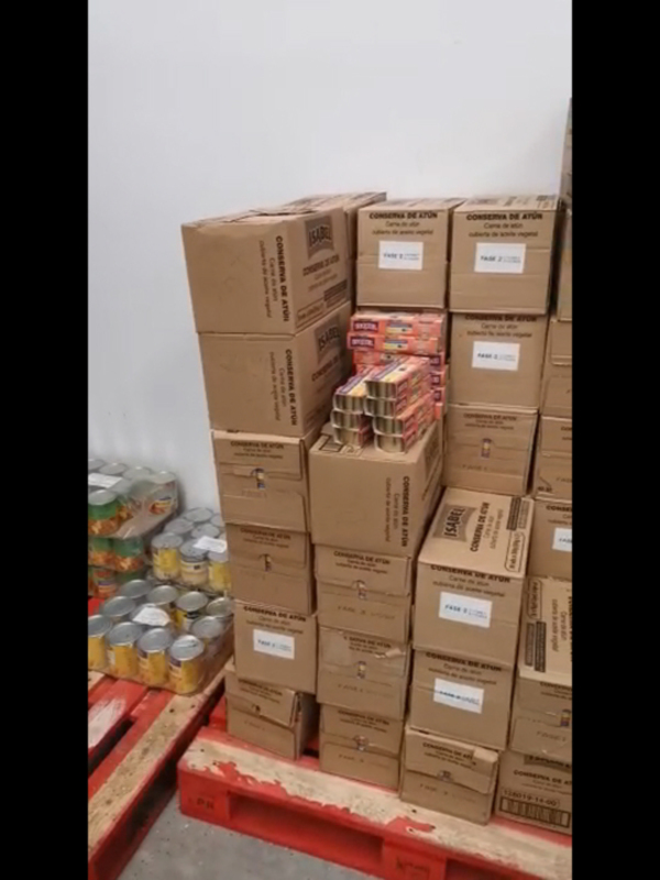 Estas imágenes muestran una gran cantidad de cajas y palets llenos de productos en un almacén. La mayoría de las cajas están abiertas y se pueden ver los productos dentro, que parecen ser latas de alimentos. La pared del almacén es blanca y las cajas están empiladas en altas montañas.