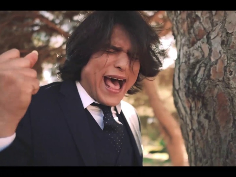 Un hombre en traje negro con corbata blanca se encuentra detrás de una rama de árbol, apuntando con su mano derecha. La escena parece ser al aire libre en un entorno natural con vegetación y luz suave.