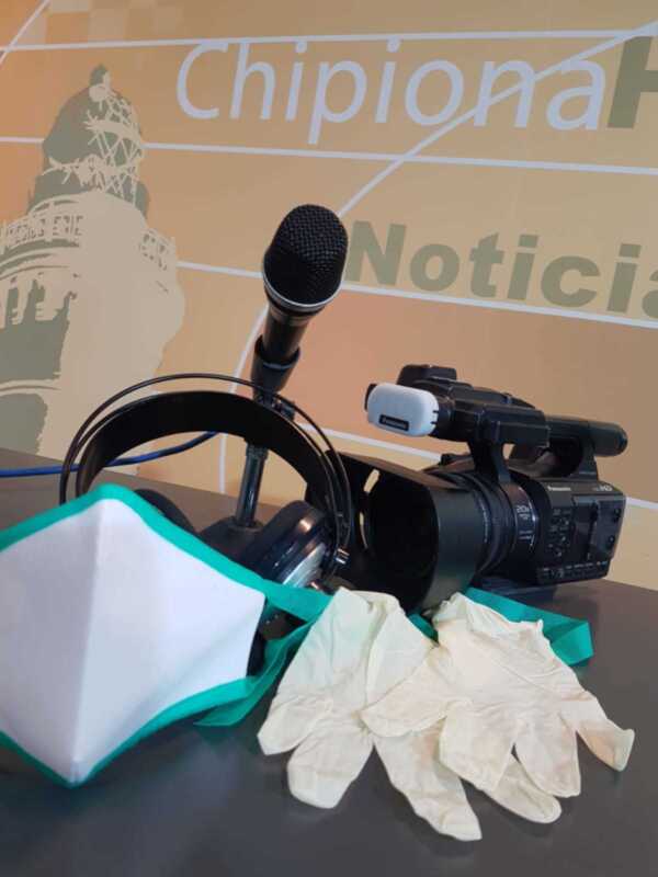 Una cámara de video y un micrófono están colocados en una mesa, con guantes desechables a su lado. En el fondo se puede ver un cartel con la palabra "Chippional" y otro con "Noticias".