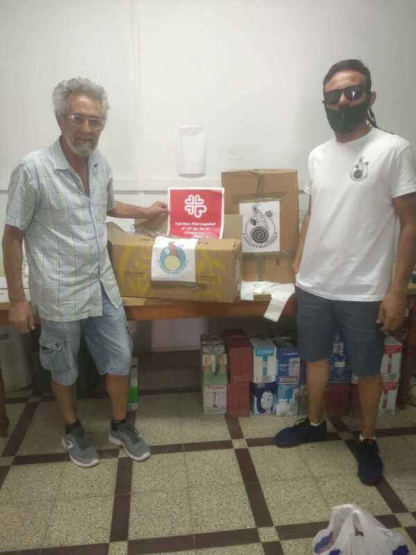 Dos hombres posan junto a cajas de donación, uno con una camiseta blanca y el otro con un jersey a cuadros. La escena se realiza en una oficina o sala de espera con un fondo blanco y azulejos.