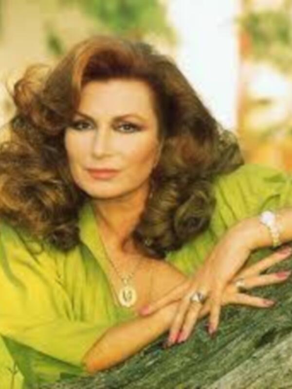 Una mujer con cabello largo y ondulado, vistiendo una camisa verde brillante. La imagen muestra detalles como un anillo en la mano y una pulsera, con un fondo que parece ser una naturaleza verde.