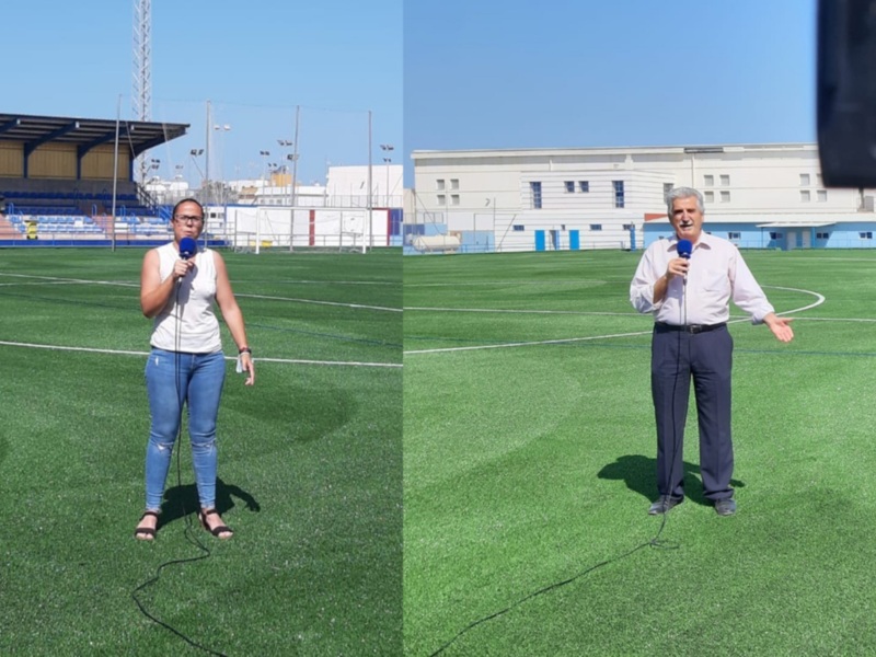 Dos personas en un campo de fútbol, una con un micrófono y otra con un auricular, ambos en actitud de hablar. Fondo: estadio con gradas vacías y edificio blanco al fondo.