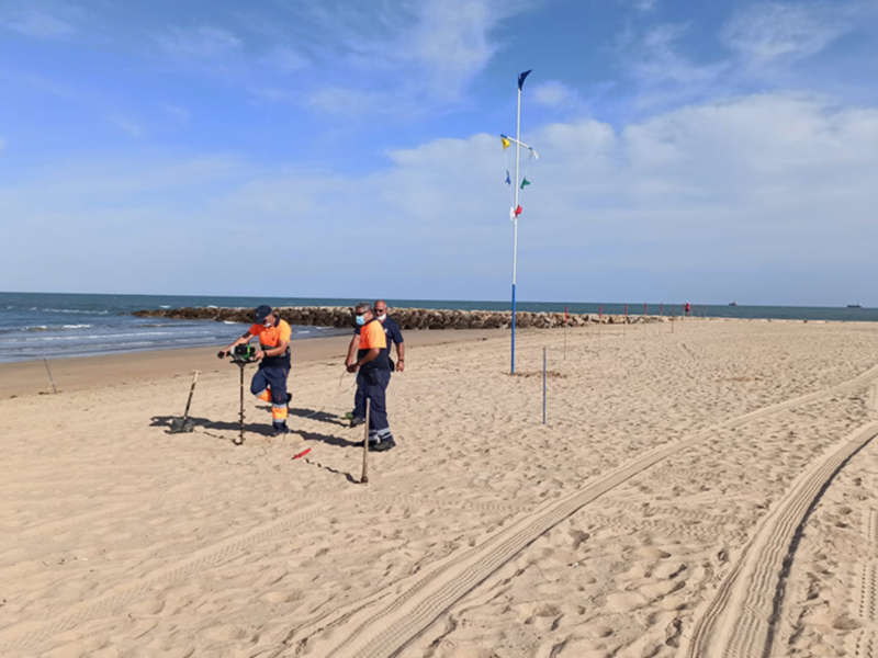 Se observan dos personas en una playa, cada uno con un equipo de metal detector. La persona a la izquierda está usando un detector de metales y una varilla para explorar el suelo, mientras que la persona a la derecha parece estar preparando o revisando su equipo. El fondo muestra un mar tranquilo con una línea de costa y cielo despejado, lo que sugiere un día soleado.