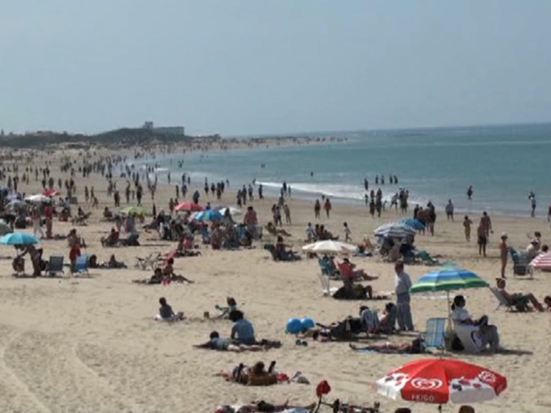 Una multitud de personas disfruta el día en la playa, con numerosas sombrillas y sillas dispuestas para descansar. La arena está llena de gente, algunas nadando en el agua cristalina y otras tomando sol bajo los umbrelas. El horizonte se ve borroso, con algunas estructuras que podrían ser edificios o monumentos a lo lejos. La atmósfera es relajante y soleada, típica de un día en la playa.