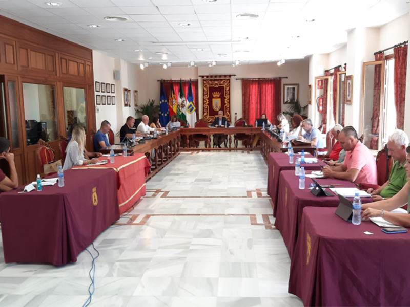 Reunión de consejo municipal en sala de plenos bien decorada con banderas y documentos en las mesas.