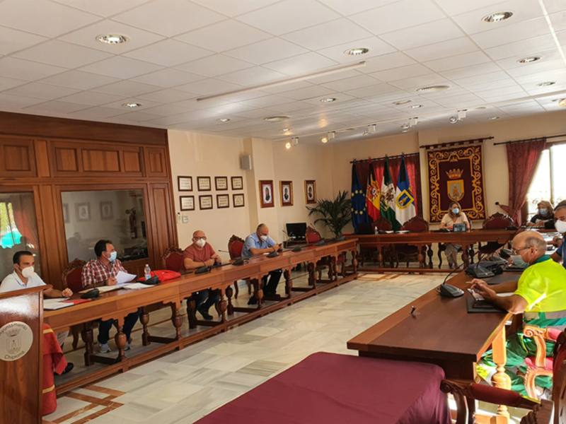Reunión formal en una sala de conferencias con varias personas sentadas en mesas, algunos portando mascarillas. La sala tiene decoración oficial y una bandera visible en el fondo.
