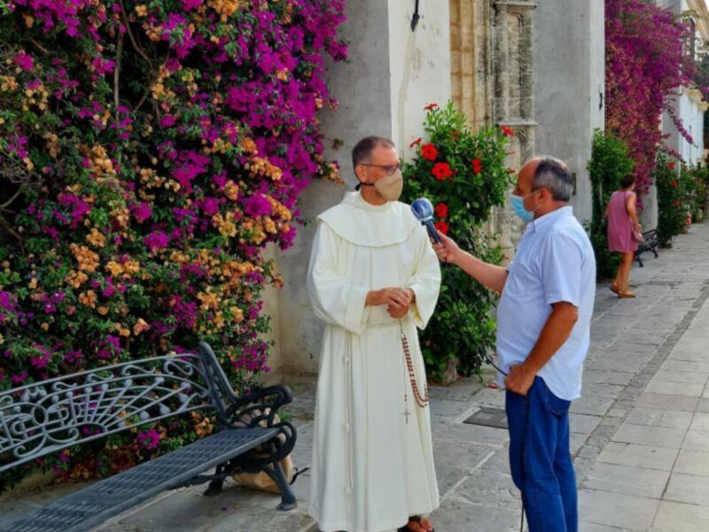 Un monje en hábito blanco habla con un hombre vestido de azul, ambos están en una calle con flores coloridas y una pared decorada.