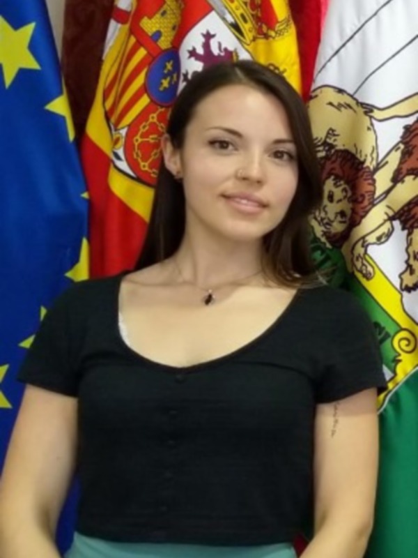 Una mujer con una camiseta negra y una falda verde posa frente a varias banderas, incluyendo la bandera de la Unión Europea y la bandera de España.