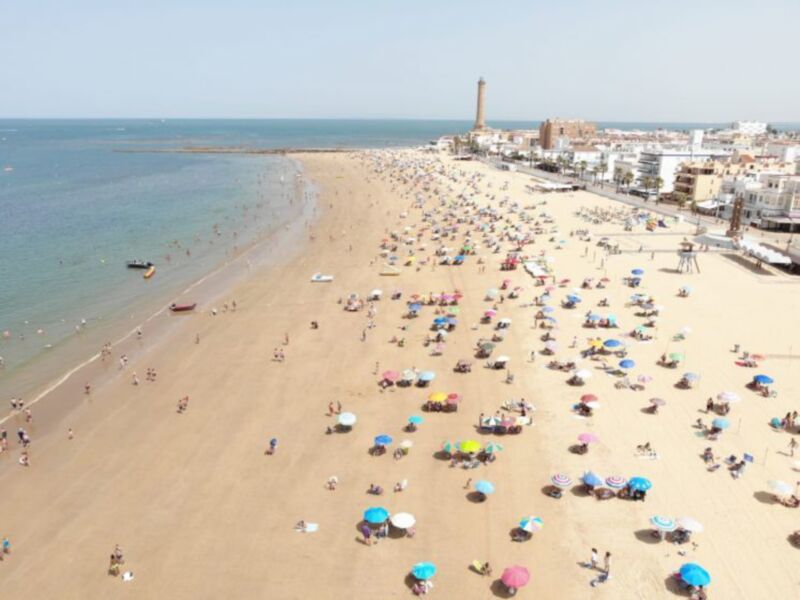 Playa de la Caleta, Cádiz, con una multitud disfrutando del sol y el mar bajo un cielo despejado.