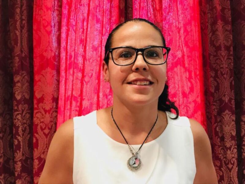 A una persona con cabello negro, peinado en dos coletas laterales. Lleva una camiseta blanca y un collar con un colgante de forma circular. El fondo es una cortina roja con detalles dorados, probablemente en un ambiente formal o ceremonial.