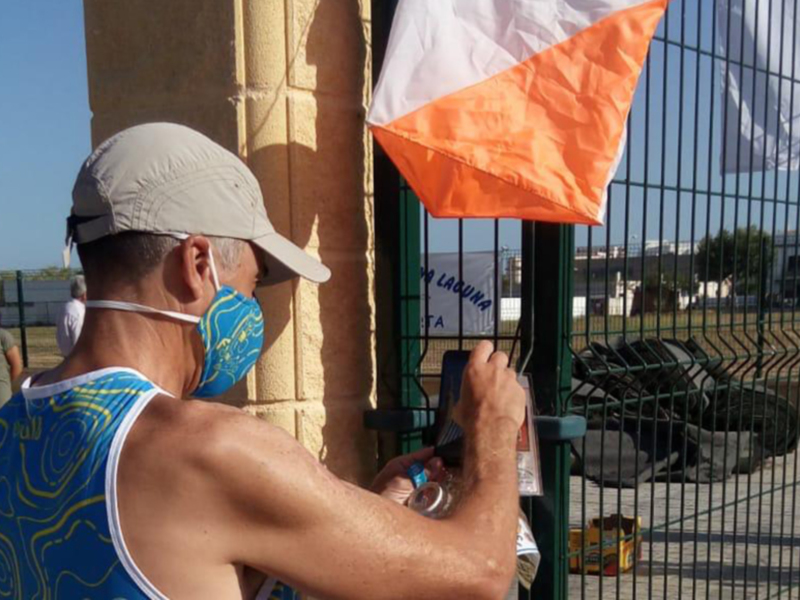 Un hombre con una gorra gris y un chaleco azul está escribiendo en una placa de metal con un marcador. Detrás de él, hay una bandera naranja y blanca ondeando en un poste de metal. La imagen se toma frente a una cerca metálica con un fondo de edificios y árboles.