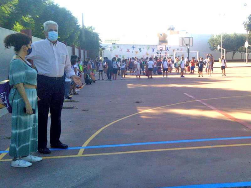 En una cancha de baloncesto, un grupo de personas observa a un niño en el centro. La imagen muestra una escena de apoyo y atención, con varios adultos y niños en el fondo. La cancha está iluminada por la luz del día, creando sombras en el suelo.