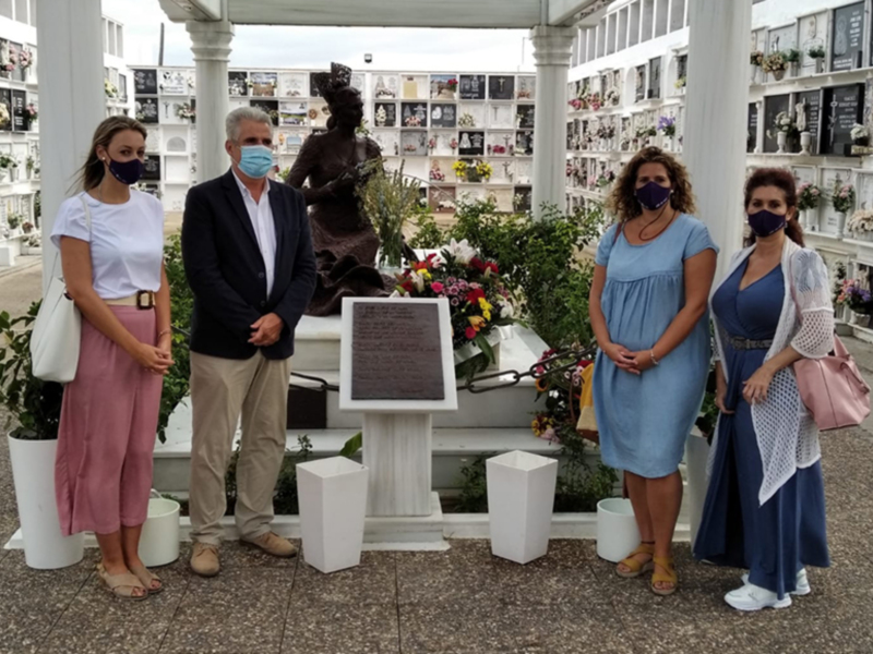 Una familia en un cementerio, con una mujer vestida de azul y otra de rosa. Todos miran hacia un monumento, con flores y una estatua en el centro. La escena está iluminada por la luz del día, con plantas y un pórtico alrededor.