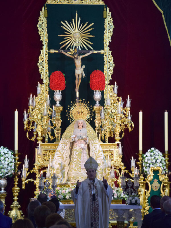 Procesión religiosa con estandarte de la Virgen y crucificado en un altar ornamental dorado.