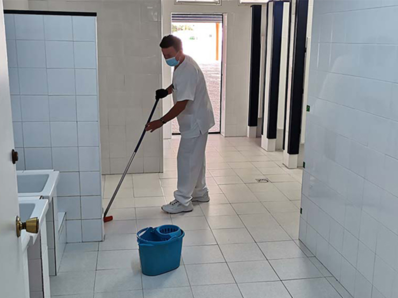 Un hombre en uniforme blanco limpiando el suelo de un baño con una escoba y una canasta azul. La pared está decorada con azulejos blancos y hay una puerta abierta al fondo.