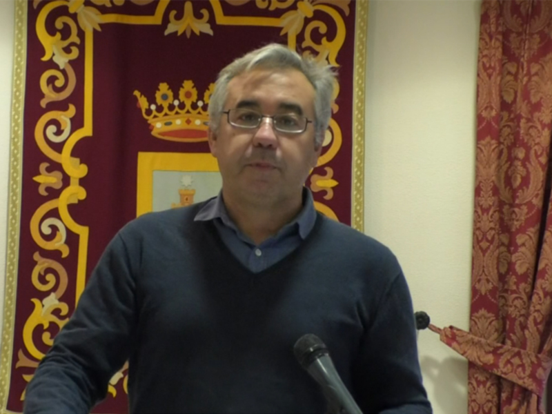 El hombre está en una sala con un escudo y una corona en el fondo, posiblemente en una oficina o sala de conferencias.