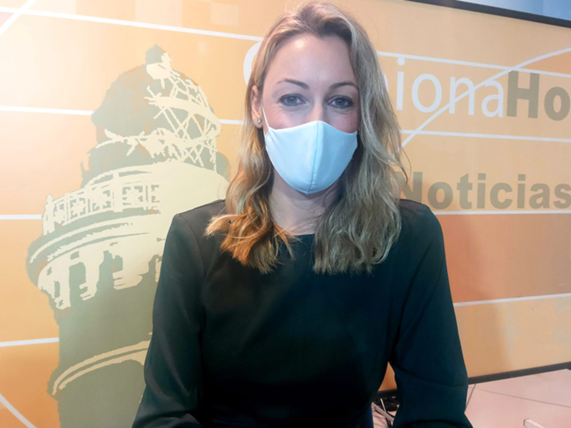 Presentadora en estudio de noticias con fondo de logo y máscara facial.