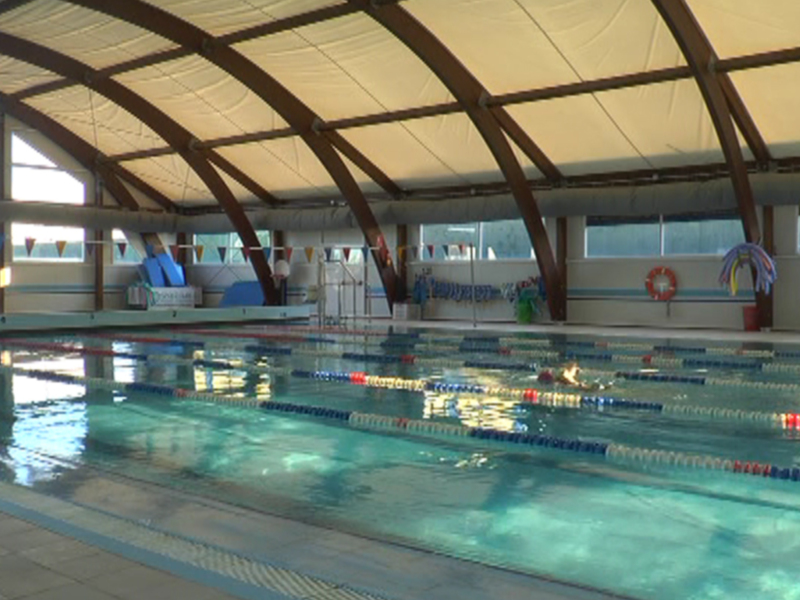Una piscina de natación interior con líneas azules y blancas en el fondo, iluminada por luz natural a través de ventanas. El techo tiene una estructura curva de madera y el suelo es azul con detalles blancos.