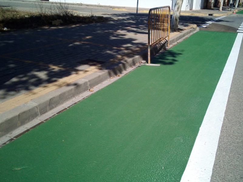 Pavimento verde en una carretera, con un carril de peatonal y un carril de coches. La imagen muestra una sección de la carretera con una línea blanca que separa los carriles. El piso es de color verde y parece estar en buen estado. Hay una barrera metálica cerca del carril de peatones, y en el fondo se pueden ver árboles y un camino.