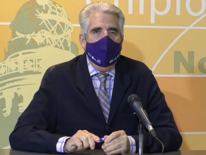 El hombre está sentado en una mesa, con un fondo amarillo y verde que parece representar el Capitolio de los Estados Unidos. Se está hablando sobre una noticia importante en un contexto político o gubernamental.