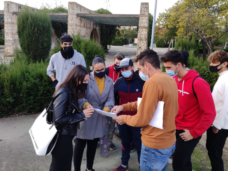 Una imagen de un grupo de personas con máscaras, posiblemente en una reunión o charla al aire libre. Algunos sujetan documentos mientras otros observan atentamente. El entorno parece ser un parque o área pública con arquitectura de piedra y vegetación alrededor.