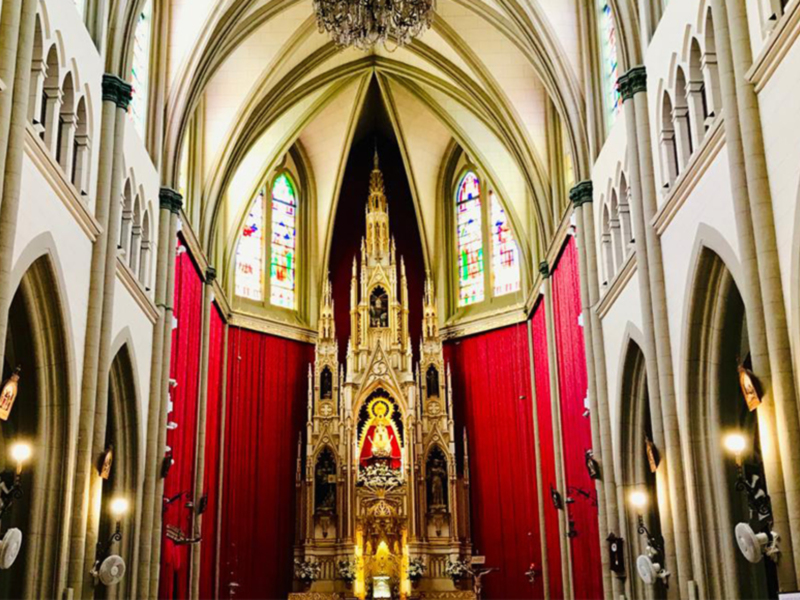Interior de una iglesia gótica con arcos altos, vitrales coloridos y un altar principal iluminado por luces doradas.