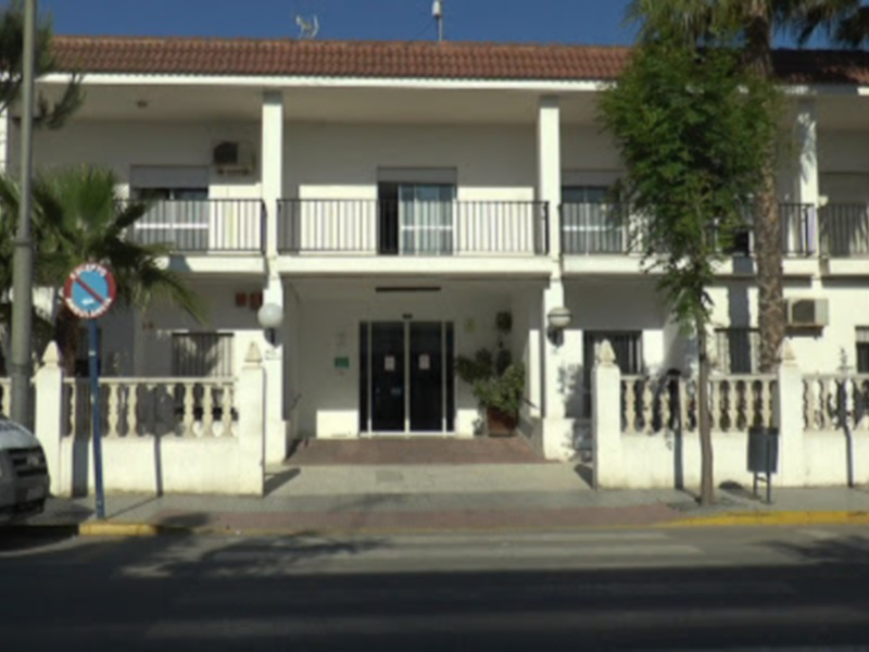 Fachada de un edificio blanco con balcones y árboles alrededor, bajo un cielo azul claro.