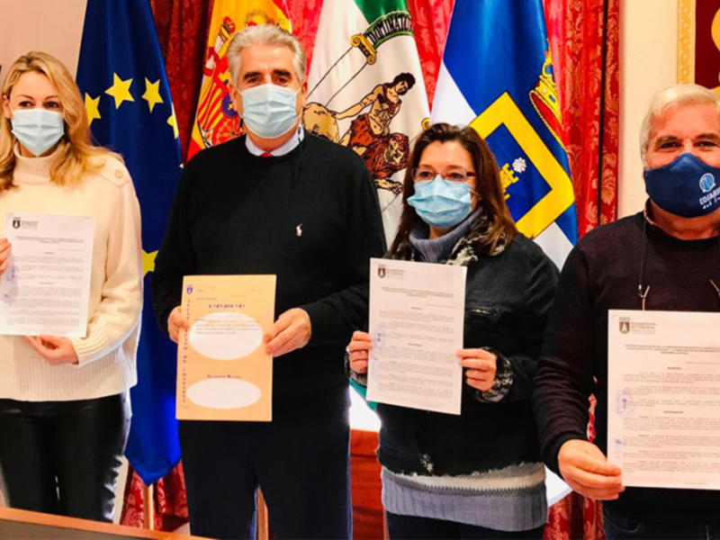 Se muestra a cuatro personas, tres hombres y una mujer, todos con mascarillas faciales, posiblemente durante la pandemia. Se pueden ver banderas de diferentes países en el fondo, lo que sugiere una reunión formal o diplomática. Los individuos están sosteniendo documentos, lo que indica un acto de negociación o firma. La escena parece ser en un entorno oficial, posiblemente una sala de conferencias o oficina gubernamental.