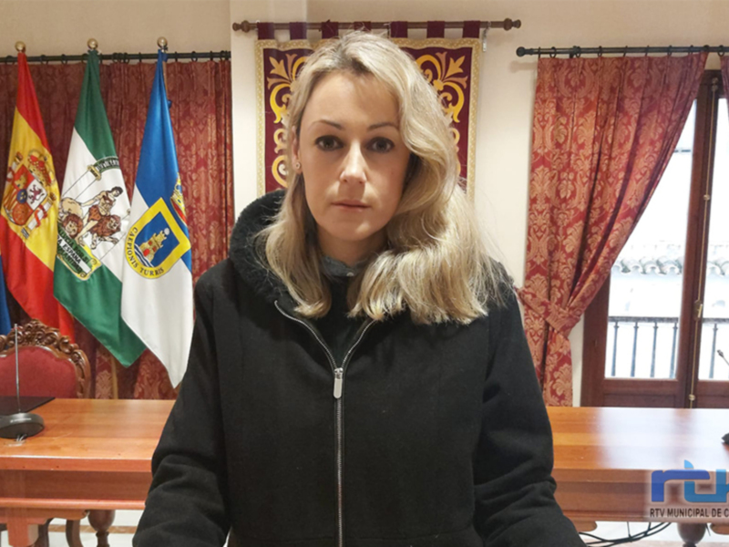 A una mujer con peluca rubia, vestida con chaqueta negra, frente a una mesa de madera. En el fondo se pueden ver varias banderas, incluyendo la de España y la de Andalucía. La escena parece ser en un entorno oficial o institucional, posiblemente una oficina gubernamental.