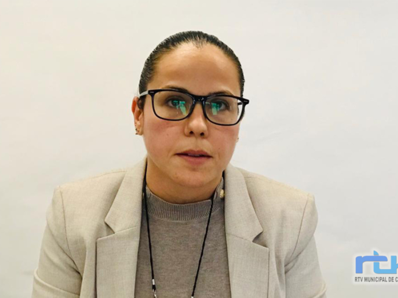 A una persona con cabello negro y rizado, llevando gafas de montura negra. Se observa una camisa gris con un blazer beige y un cuello de lana. La persona está en una habitación con paredes blancas y el logo "RTV" visible en la esquina inferior derecha.