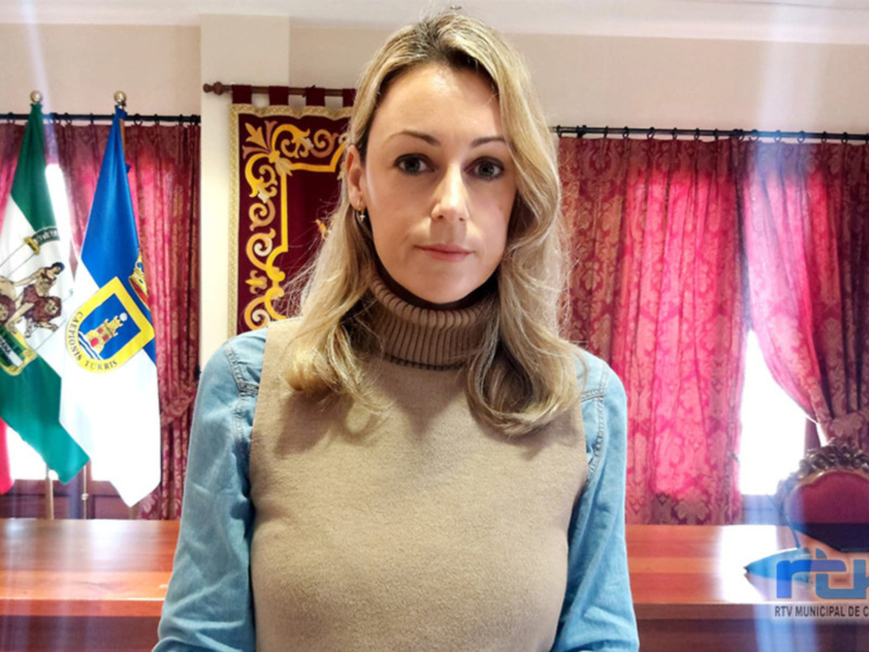 A una mujer con cabello largo y rubio, vestida con una camiseta beige y una blusa azul. El fondo incluye banderas de España, Andalucía y Murcia, junto con cortinas rojas. La escena parece ser en un entorno oficial o institucional, posiblemente una oficina municipal.