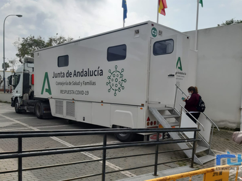 Un camión blanco de la Junta de Andalucía, con el lema "Respuesta COVID-19", está estacionado en un área de aparcamiento. La imagen muestra una persona caminando hacia el vehículo, que parece ser un centro móvil de salud. La bandera de Andalucía está claramente visible en el fondo, y hay un poste con una bandera roja y amarilla a la derecha del camión.