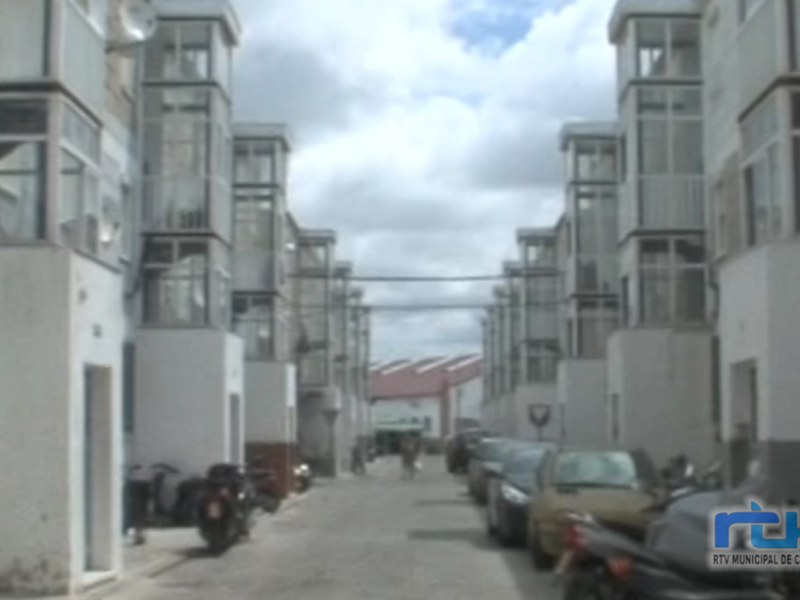 Una calle de edificios residenciales en un entorno urbano. Las casas son modernas y alineadas, con ventanas grandes que permiten una buena iluminación natural. El cielo está nublado, lo que sugiere un día con algunas nubes pero sin lluvia. En la calle hay varios vehículos estacionados, incluyendo coches y motocicletas. La perspectiva de la imagen sugiere que se está mirando hacia el final de la calle.