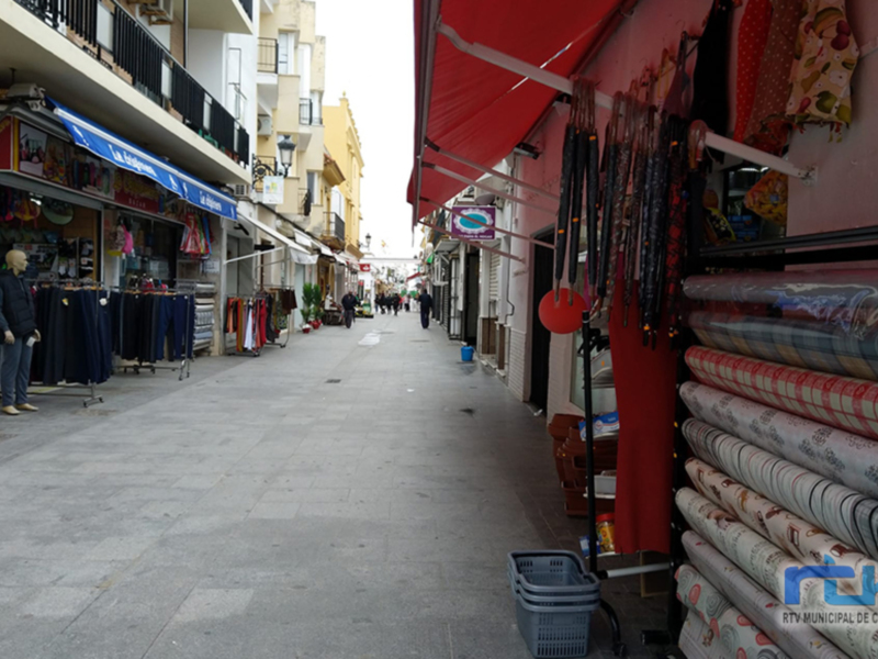 Imagen de una calle comercial con tiendas y estantes llenos de productos. El cielo está nublado, lo que sugiere un clima frío o invernal. La calle está desierta, con pocos pasajeros y la luz del día es tenue.
