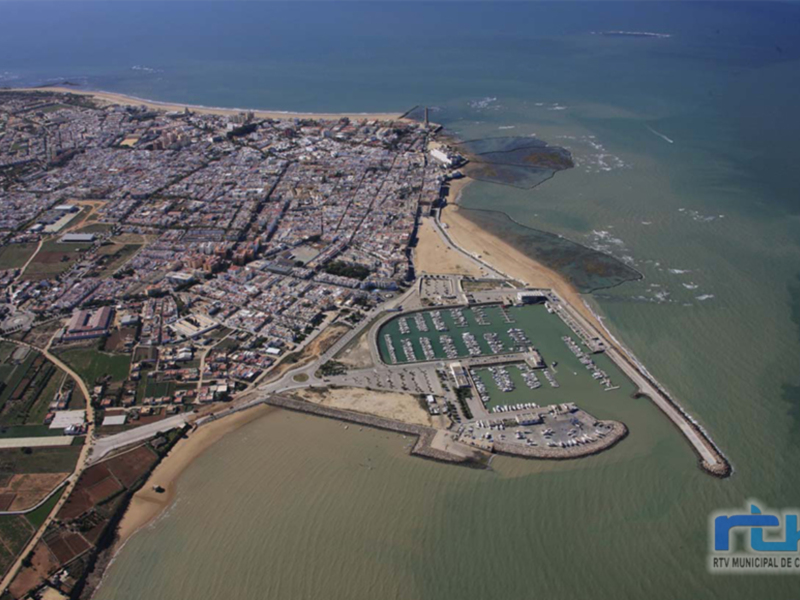Imagen aérea de Chipiona, una localidad costera en Cádiz. Se observa la ciudad con sus edificios, el puerto y la playa. La imagen muestra la arquitectura urbana y la costa con aguas turquesas.