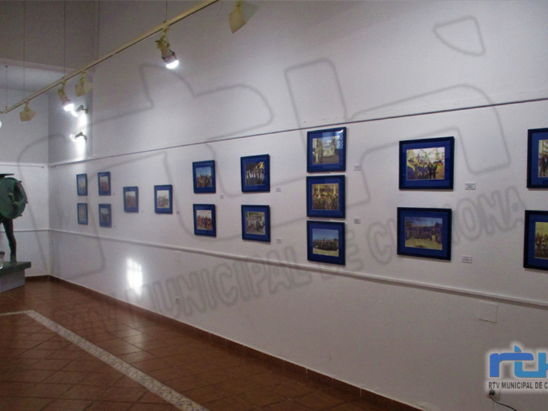 Una sala de exposiciones con varias fotos enmarcadas colgando en una pared blanca. La sala tiene suelo de baldosas marrones y luces colgantes en la parte superior. La imagen incluye un nombre de la ciudad, "Chihuahua", y una marca de la empresa.