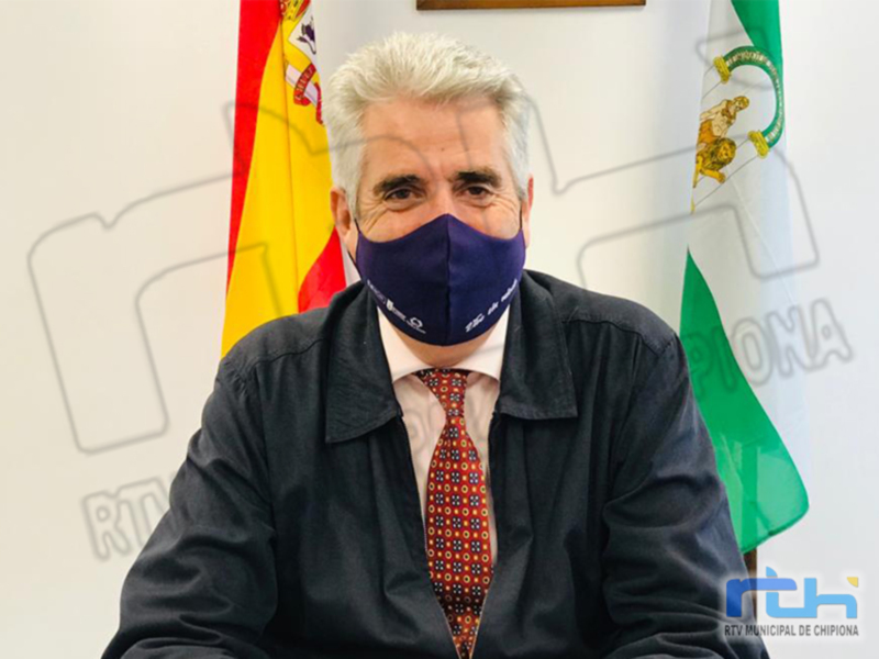 Un hombre con cabello blanco y barba, usando una máscara facial azul, se encuentra en un entorno formal con banderas española y andaluza al fondo.