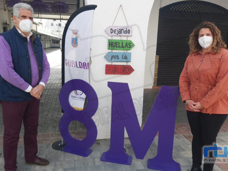 Dos personas posan junto a un gran número 8M, con carteles que mencionan "Dejando huellas por la igualdad".