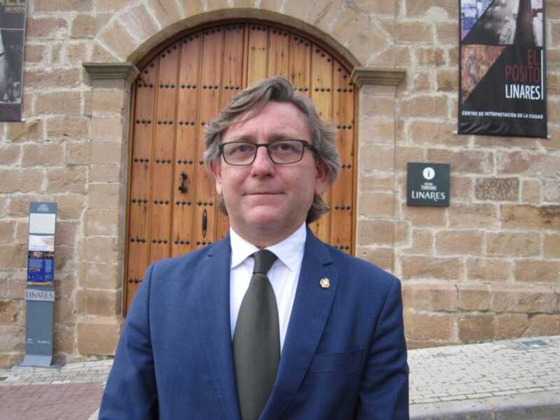 El hombre está vestido con una chaqueta azul y un corbata negra, frente a una pared de ladrillo con una puerta de madera y dos carteles en la parte superior.