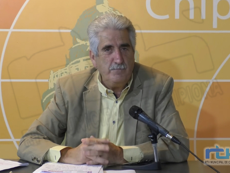 El hombre está sentado frente a un fondo con el logo de la municipalidad de Chipiona, hablando en una conferencia o reunión.