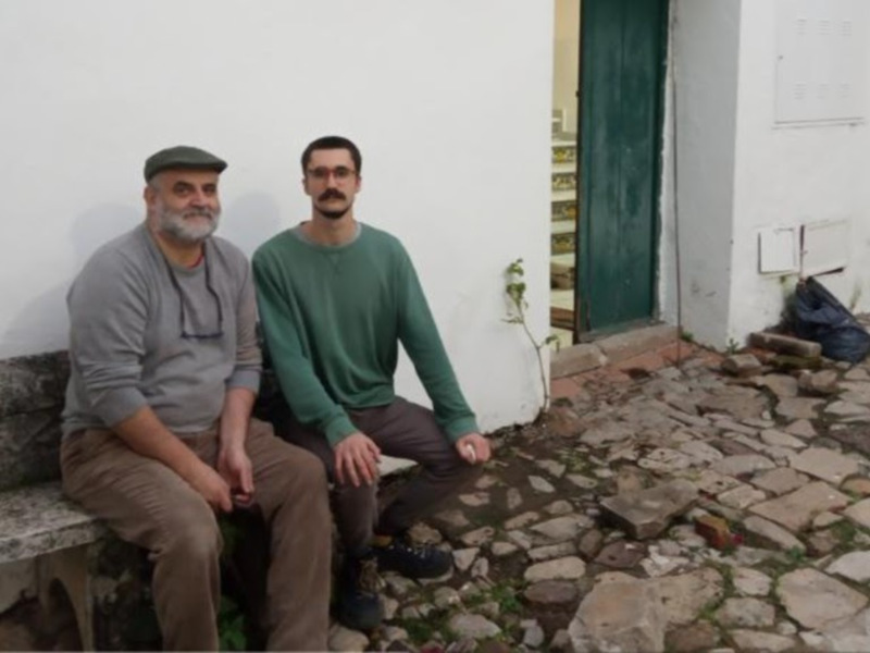 Dos personas sentadas en una banca de piedra frente a una pared blanca y una puerta verde, en un entorno rural con adoquines y una bolsa de basura en el suelo.
