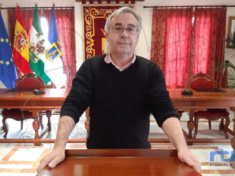 Un hombre con gafas y una camiseta negra se encuentra sentado en un escritorio frente a varias banderas, incluyendo la de España. La imagen está tomada en un entorno formal con cortinas rojas y una mesa de conferencia.