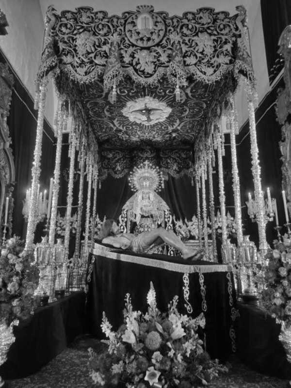 Una imagen en blanco y negro muestra una figura vestida de blanco, posiblemente representando a la Virgen María, sentada sobre un trono elaborado. El trono está adornado con flores y luces, creando un ambiente festivo y religioso. La figura está rodeada por una atmósfera de solemnidad y reverencia, típica de la cultura católica.