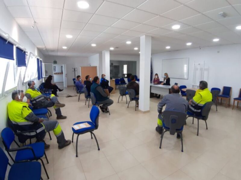Un grupo de personas en uniforme amarillo-verde se reúne en una sala con paredes blancas y suelo de baldosas. La sala tiene sillas azules, una mesa con documentos y un panel blanco en la pared. Algunos asistentes están sentados, mientras otros se levantan para hablar. La iluminación es natural y la atmósfera parece ser de trabajo o capacitación.
