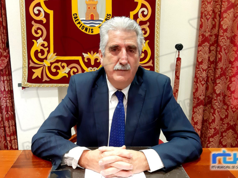 Un hombre en traje azul y corbata azul se sienta frente a una bandera con un escudo en el fondo. La imagen parece ser de una oficina o sala formal, con cortinas rojas y doradas.