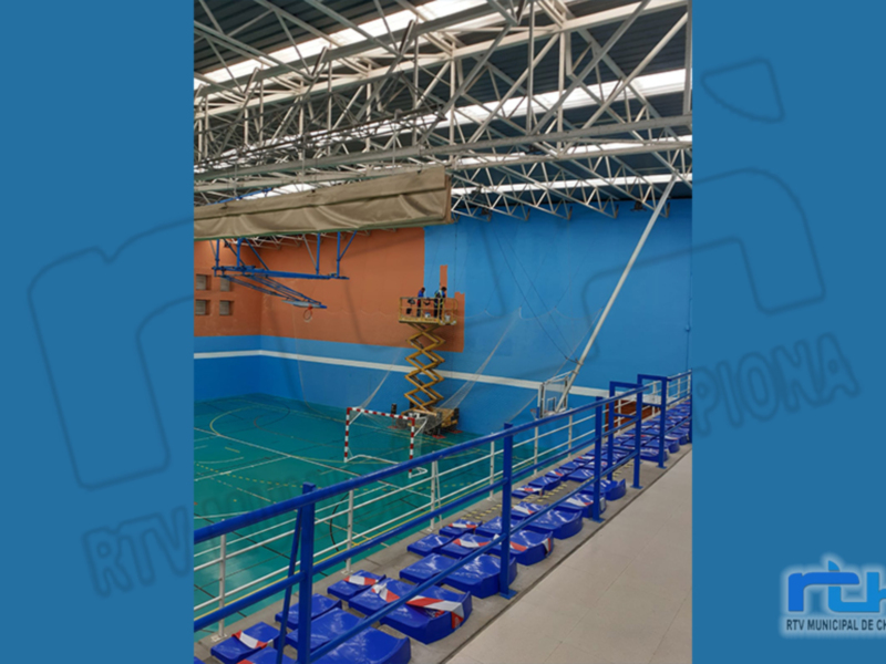 Un gimnasio con canchas de baloncesto y voleibol, techos altos con estructuras metálicas, y una grúa en funcionamiento.