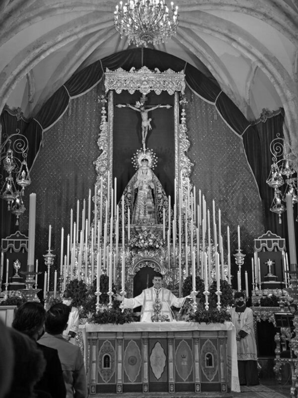 Sacerdote oficiando una misa en un altar ornado con velas y una imagen religiosa, en un ambiente de iglesia histórica.